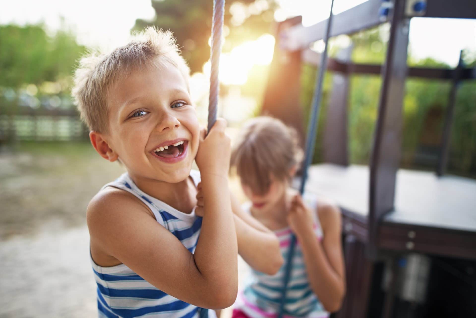 Two kids standing together outdoors, laughing and having fun. They are dressed casually, with one child in the foreground wearing a striped shirt and holding onto a playground structure.